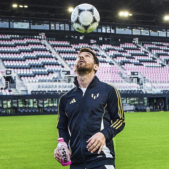 Leo Messi juggling a soccer ball on a sports field with Más by Messi in his hand