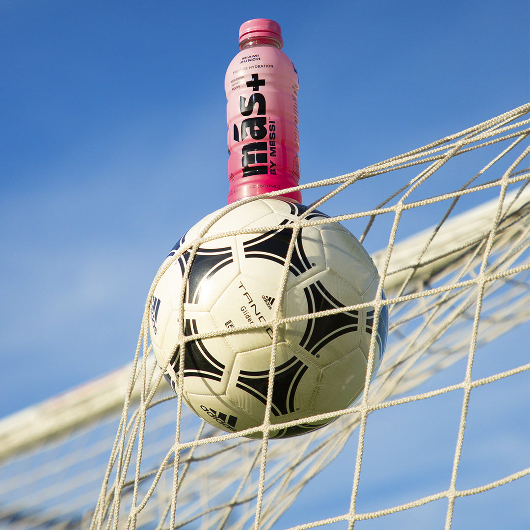 A pink Más+ by Messi hydration bottle balanced on top of a football caught in the goal net against a clear blue sky.