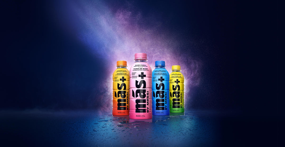 Four vibrant bottles of Más+ by Messi flavoured drinks are arranged in a row against a dark blue, misty background. Each bottle has a different coloured label: orange, pink, blue, and yellow and a volume of 500 mL displayed on each.