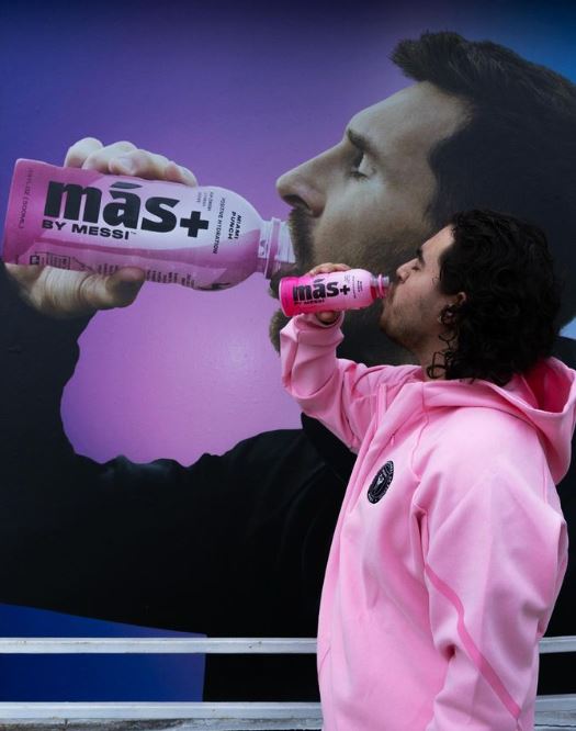 A person in a pink hoodie drinking Más+ by Messi next to an advertisement where Leo Messi is doing the same.