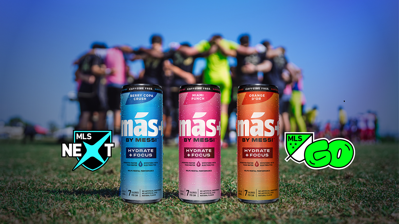 Three cans of Más+ by Messi drink (blue, pink, orange) on grass in front of blurry soccer players and MLS NEXT and GO logos.
