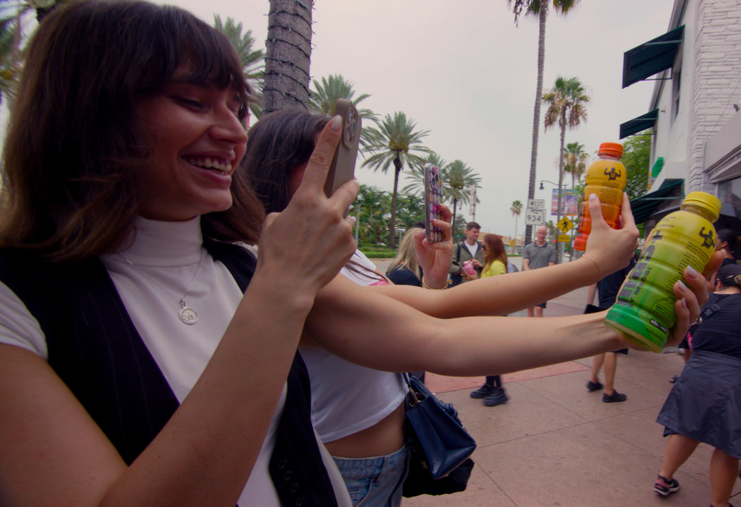 Two girls holding a bottle of Más+ by Messi drink while taking photos.