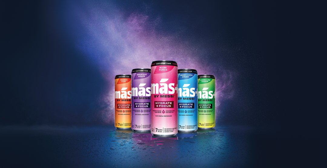 Five cans of Más+ by Messi drink are lined up on a dark, wet surface under moody blue and purple lighting.